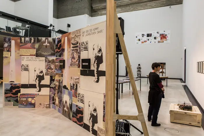 Ein großer Ausstellungsraum mit einem Holzschirm im Vordergrund, auf dem verschiedene Poster hängen. 